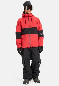 Rote und schwarze Skijacke mit kariertem Muster, Reißverschluss, verstellbarer Kapuze, getragen mit schwarzer Schneehose und Handschuhen. Große Skibrille.
