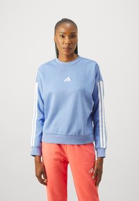 Ljusblå sweatshirt med vitt adidas-logo på bröstet och tre vita ränder längs ärmarna, ihopparad med korallfärgade sweatpants.