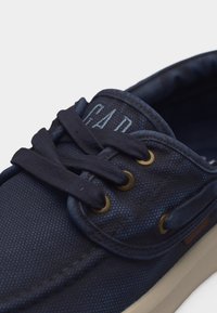 Chaussure bateau en toile bleu marine avec lacets noirs, œillets en laiton et semelle en caoutchouc beige. Logo partiel visible sur la languette rembourrée.