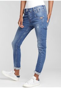 Persoon in opgerolde blauwe denim jeans met lichte slijtage en witte sneakers, staande tegen een effen lichte achtergrond.