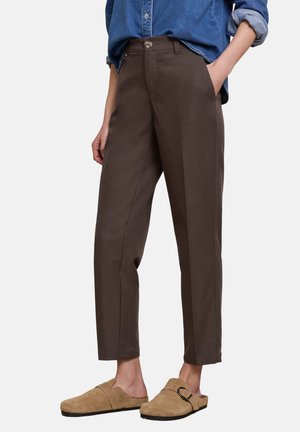 Osoba ubrana w ciemnobrązowe zwężane spodnie, niebieską podwiniętą koszulę dżinsową i beżowe wsuwane buty z ozdobną klamrą.