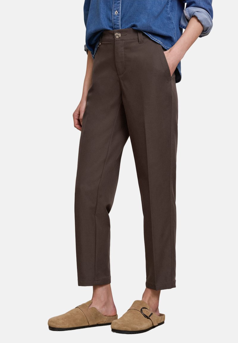 Persona vistiendo pantalones entallados marrón oscuro, camisa de mezclilla azul remangada y zapatos beige sin cordones con detalle de hebilla.