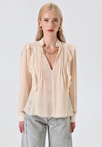 Blusa beige con maniche lunghe, scollo a V, dettaglio a volant sul davanti e polsini con bottoni. Indossata con jeans grigi chiari e morbidi.