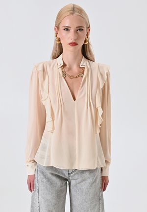 Blusa beige con maniche lunghe, scollo a V, dettaglio a volant sul davanti e polsini con bottoni. Indossata con jeans grigi chiari e morbidi.