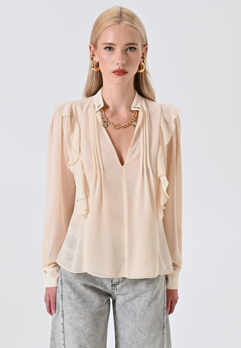 Blusa beige con maniche lunghe, scollo a V, dettaglio a volant sul davanti e polsini con bottoni. Indossata con jeans grigi chiari e morbidi.