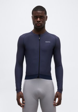 Άνδρας που φοράει μπλε σκούρο ποδηλατικό ζακέτα με φερμουάρ και μακριά μανίκια, και ανοιχτό γκρι επενδυμένο ποδηλατικό σορτς, στέκεται μπροστά σε απλό φόντο.