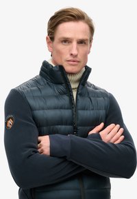 Ciemnoniebieska kurtka puchowa w stylu z wysokim kołnierzem, zapinana na zamek, z kontrastującymi granatowymi rękawami z polaru. Posiada naszywkę marki na lewym ramieniu.