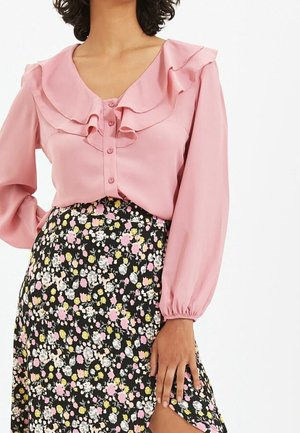 Vrouw draagt een roze blouse met ruches aan de kraag en knopen, gecombineerd met een zwarte bloemenrok met een hoge split aan de dij.