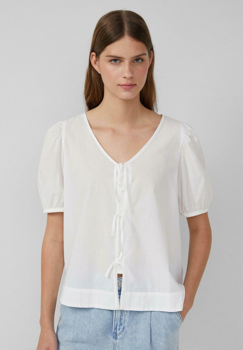 Blusa bianca a maniche corte con scollatura a V, dettagli con fiocco frontale e maniche a sbuffo. Realizzata in un tessuto leggero con una vestibilità ampia.