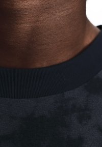 Gros plan sur le cou d'une personne à la peau foncée portant une chemise à col côtelé foncé sur fond blanc.