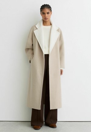 Béžový nadrozmerný vlnený kabát s bielym golierom, širokými rukávmi a bočnými vreckami, doplnený krémovým svetrom a hnedými nohavicami.