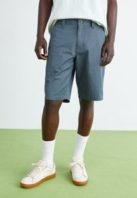 Pantaloni scurți gri dintr-un material cu o textură netedă, cu închidere prin buton și fermoar. Asortați cu șosete albe și adidași crem.