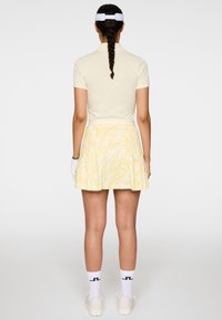 Femme aux cheveux tressés portant un polo jaune pâle, une jupe jaune à motifs, des gants de sport blancs, des chaussettes blanches et des baskets blanches, debout de dos.