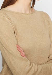 Beige gebreide trui met een gestructureerde afwerking, ronde halslijn en lange mouwen, met een ontspannen pasvorm en geribde accenten bij de mouwen.