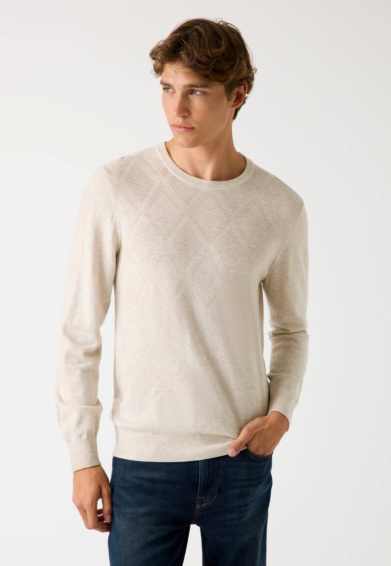 Giovane uomo con un maglione di cotone chiaro a motivo a rombi e jeans blu scuro, in piedi con una mano in tasca, mentre guarda di lato.