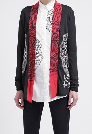Person wearing a black cardigan with red and white abstract patterns over a white button-up shirt and black pants.