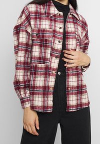 Czerwona i biała koszula flanelowa w kratę z zapięciem na guziki, z kołnierzykiem i dwoma kieszeniami na piersi, noszona na czarnej bluzce i zestawiona z ciemnymi spodniami.