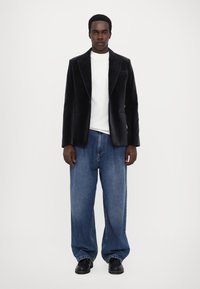 Juodas aksominis švarkas virš balto marškinėlio, derinamas su laisvais mėlynais džinsais ir juodais batais. Lygus paviršius ir struktūrizuotas dizainas.