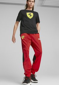 Camiseta negra con el logotipo amarillo de Ferrari, pantalones deportivos rojos con acentos negros, puños elásticos y logotipo, combinados con zapatillas negras.