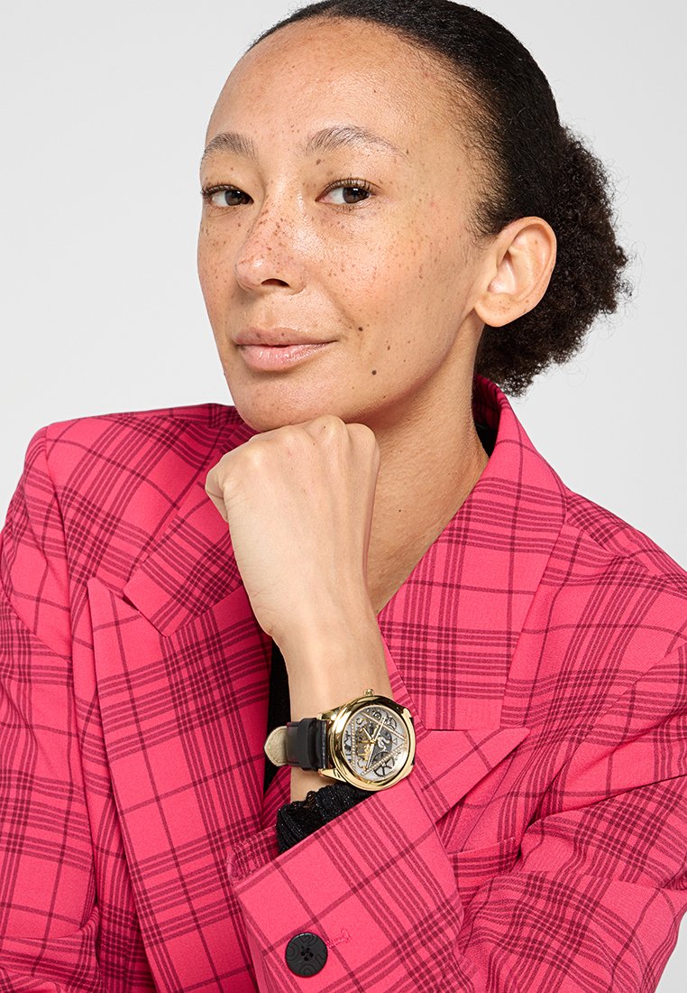 Orologio da polso dorato con cinturino in pelle nera, il quadrante mostra dettagli intricati. Indossato con un blazer a quadri rosa, caratterizzato da un forte motivo geometrico.