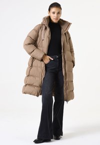 Abrigo marrón largo y voluminoso con capucha y cremallera, de material texturizado, combinado con jeans negros de corte acampanado y zapatos oscuros.