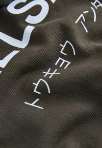 Tissu en coton kaki avec un texte blanc proéminent et une texture lisse. Le texte présente à la fois des caractères latins et des caractères stylisés de tailles variées.