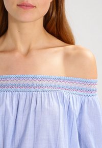 Helles blaues Off-Shoulder-Oberteil mit elastischem, gerafftem Ausschnitt, das mehrfarbige Stickereien und einen fließenden Stofftextur aufweist.