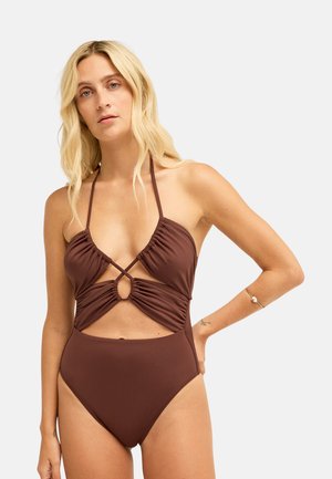 Femme aux longs cheveux blonds vêtue d’un maillot de bain une pièce marron avec découpes et bretelles halter, debout devant un fond uni.