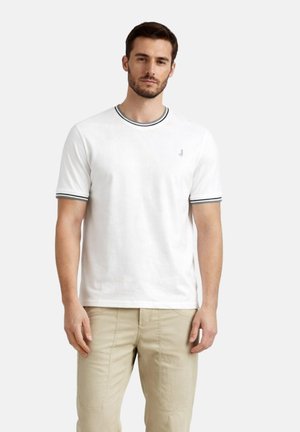 Uomo che indossa una T-shirt bianca a maniche corte con bordi neri su colletto e maniche, abbinata a pantaloni beige, in piedi davanti a uno sfondo semplice.