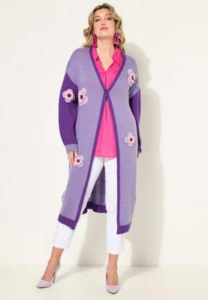 Langer lila Cardigan mit floralen Häkelverzierungen, Knopfverschluss und gerippten Bündchen, kombiniert mit einem pinken Oberteil und weißen Hosen.