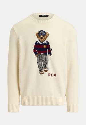 Homme portant des lunettes de soleil, pull crème avec un motif d'ours en peluche, pantalon plissé gris, chaussures blanches, debout les mains dans les poches sur fond uni.