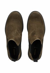 Zapatos de ante marrón tipo slip-on con paneles laterales elásticos, punta redonda y una suela de goma texturizada. La marca es visible en el interior del zapato.