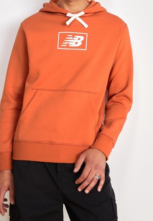 Oranje hoodie gemaakt van zacht materiaal, met een voorzak, ribgebreide manchetten en een wit merklogo in een rechthoekige omranding op de borst.