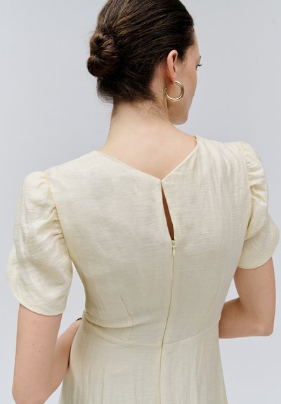 Frau mit dunklem Haar im Dutt, die goldene Creolen trägt und ein cremefarbenes, strukturiertes Kurzarmkleid mit Reißverschluss hinten und kleinem Schlitzdetail trägt.