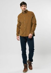 Brun fløjlskjorte med knapper og et lille logo, parret med mørkeblå jeans og brune læderstøvler.