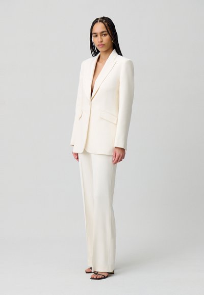 Conjunto de traje de blazer y pantalones en marfil, con un escote en V profundo, corte entallado, textura suave y diseño minimalista. Sandalias negras con tiras.