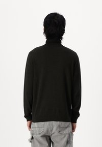 Mężczyzna z ciemnymi włosami, ubrany w czarny sweter z wysokim kołnierzem i jasnoszare spodnie cargo, stoi tyłem na białym tle.