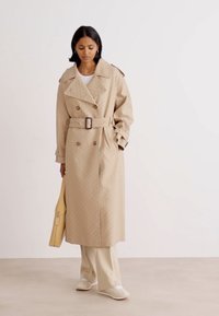 Manteau beige avec un design croisé, taille ceinturée, col oversized et texture subtile. Assorti à un pantalon et des chaussures de couleur crème.