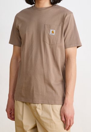 Personne portant un t-shirt marron à manches courtes avec une poche avant et un pantalon beige, debout devant un fond clair uni.