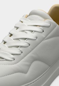 Λευκό δερμάτινο αθλητικό παπούτσι με λευκά υφασμάτινα κορδόνια δεμένα πάνω σε λείο μαλακό γλώσσα και ραμμένες λεπτομέρειες στο πλάι, πάνω σε απλό φόντο.