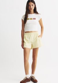 Valkoinen, istuva crop-top, jossa on värikäs graafinen suunnittelu; vaaleankeltaiset raidalliset shortsit, joissa on joustava vyötärö ja sivutaskut; ruskeat lenkkarit.