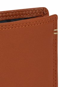 Angolo di un portafoglio in pelle marrone che mostra una texture liscia, cuciture a contrasto e una leggera lucentezza. L'interno rivela un rivestimento in tessuto scuro.