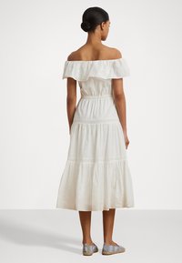 Bela off-shoulder obleka z nabranim ovratnikom, elastičnim pasom, vezenimi detajli in slojevskim krilom. Nosi se z prugastimi espadrilami.