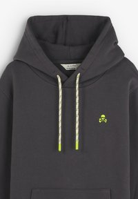 Sudadera gris oscura con un bolsillo frontal, con cordones en verde neón y un pequeño emblema de calavera en el pecho. Material de algodón.