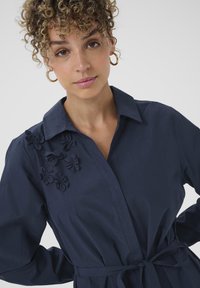 Robe chemise bleu marine avec appliqué floral sur l'épaule gauche, col classique, manches longues et ceinture à nouer. Texture de tissu lisse.
