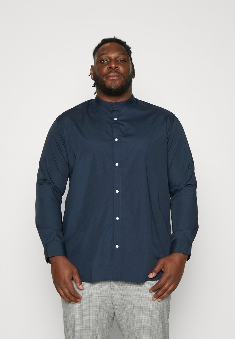 Navy blue, long-sleeve button-up shirt with a stand collar and white buttons, featuring a smooth texture. Worn with checkered gray trousers.