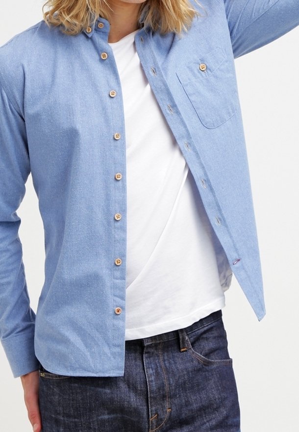 Personne portant une chemise bleu clair déboutonnée par-dessus un t-shirt blanc et un jean bleu foncé, levant légèrement un bras.