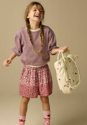 Jong meisje met gevlochten haar lacht terwijl ze een crèmekleurige rugzak met hartpatronen vasthoudt, gekleed in een gestreepte trui en roze bloemenbroekje.