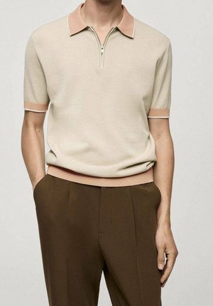 Homme portant un polo en maille beige à manches courtes avec fermeture éclair et un pantalon marron, debout les mains dans les poches sur un fond uni.