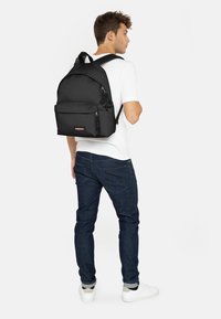 Mochila negra de nailon con forma redondeada, correas de hombro acolchadas, bolsillo frontal y etiqueta de marca. Textura suave, diseño minimalista.
